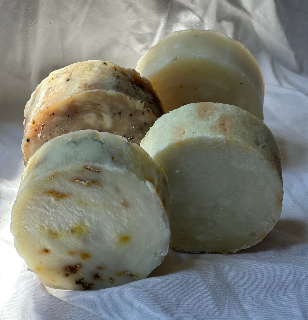 Four round, sliced bars of handmade soap with textured, speckled organic-looking tops, arranged on a wrinkled white fabric background.